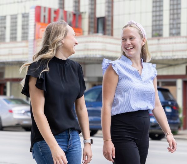 Two women smile at each other as they walk on a sidewalk together.