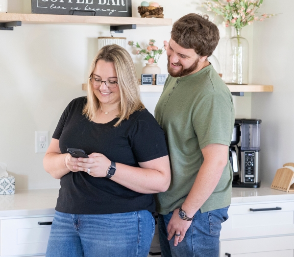 A couple stands together in a kitchen as the husband watches his wife use digital banking on her phone.