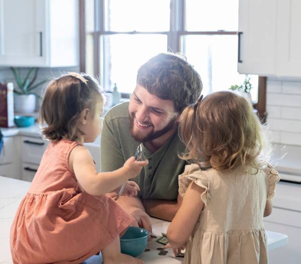 A dad smiles at his two daughters as they play with money on a countertop.