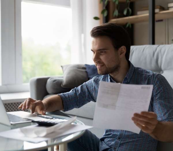 A man sits on the floor of his living room against his couch as he views documents and works on his laptop.