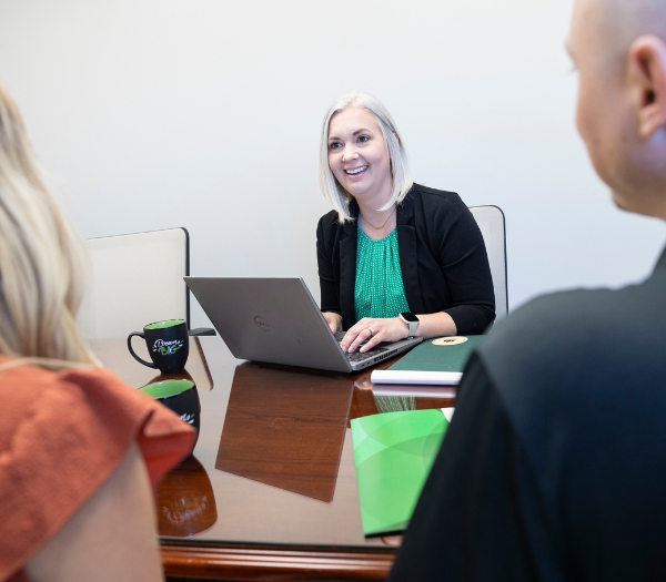 A female mortgage lender sits at a conference table while conversating with a young couple and typing on her laptop.