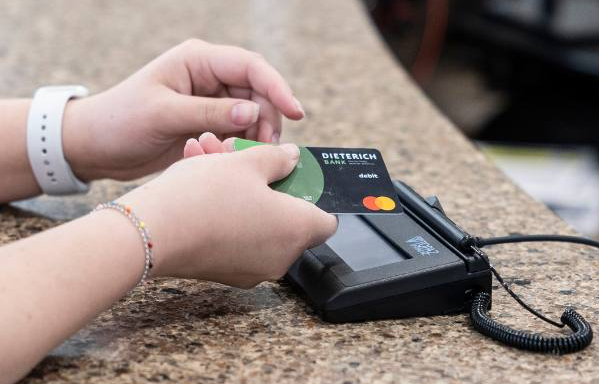 A hand guides a debit card towards a terminal sitting on a desk.