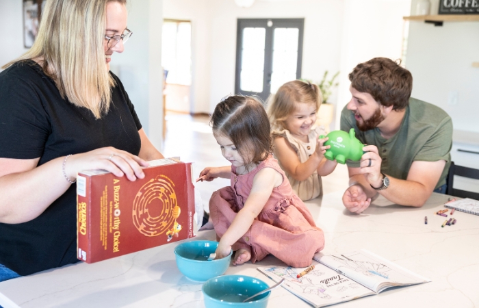 A dad plays with his daughter and her piggybank, while a mom pours cereal for another daughter sitting at a counter.